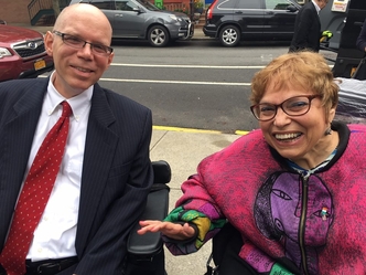 Dr Chris Rosa wearing glasses, a dark colored suit and a red tie in his wheelchair and Judy Heumann wearing glasses and wearing a purple cardigan with a bright pattern on it sitting in her wheelchair to the right of him. They are outside on a city sidewalk.