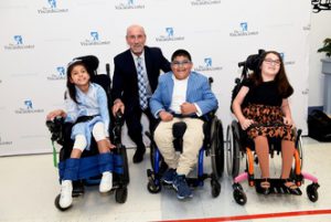 Former NY Islander Bobby Nystrom wearing a suit crouched down behind three elementary school age students - two female and one male – sitting in wheelchairs in front of a The Viscardi Center logo wall.