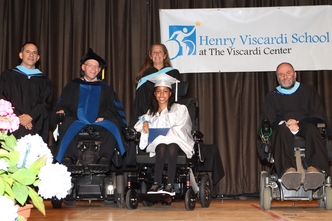 Female student in white cap and gown receiving diploma on a stage with a Henry Viscardi School at The Viscardi Center banner as a backdrop. Several adults in black graduation gowns are behind and next to her.