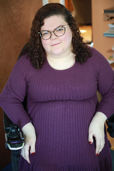 Caucasian woman with brown curly hair, wearing glasses and a purple long-sleeved sweater dress, sitting in a wheelchair.
